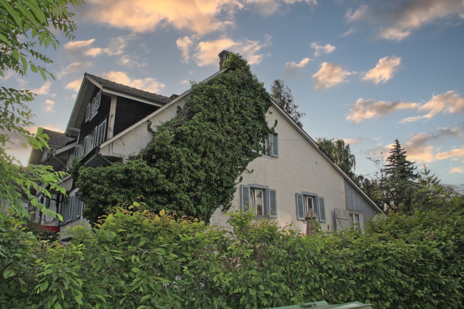 Two-story house with a gabled roof, surrounded by lush greenery and vegetation. The house has shuttered windows and a balcony or terrace visible on the upper level. The exterior walls are painted a light color, and the roof appears to be made of tiles or
