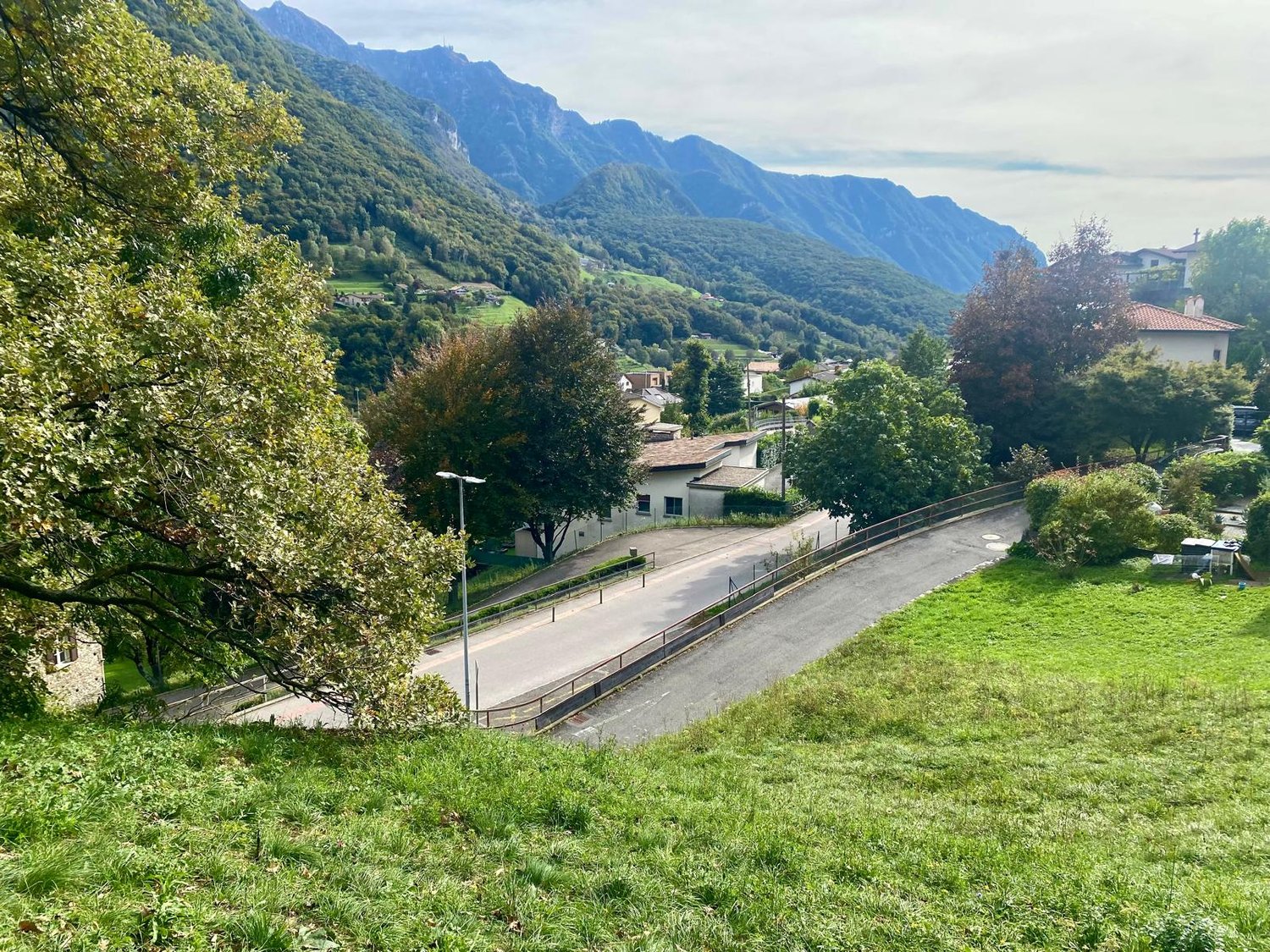 The image shows a scenic mountain landscape with a small village nestled in the valley. The scene features lush green foliage, including trees and grassy areas, as well as a winding road leading through the village. The mountains in the background are tal