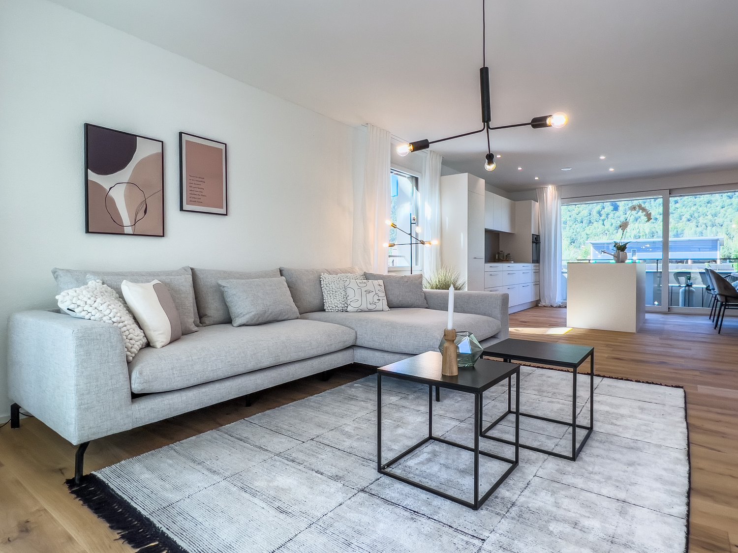 Spacious living room with a large gray sectional sofa, black coffee tables, abstract wall art, and a modern lighting fixture. The room has large windows providing a view of the outdoor area.