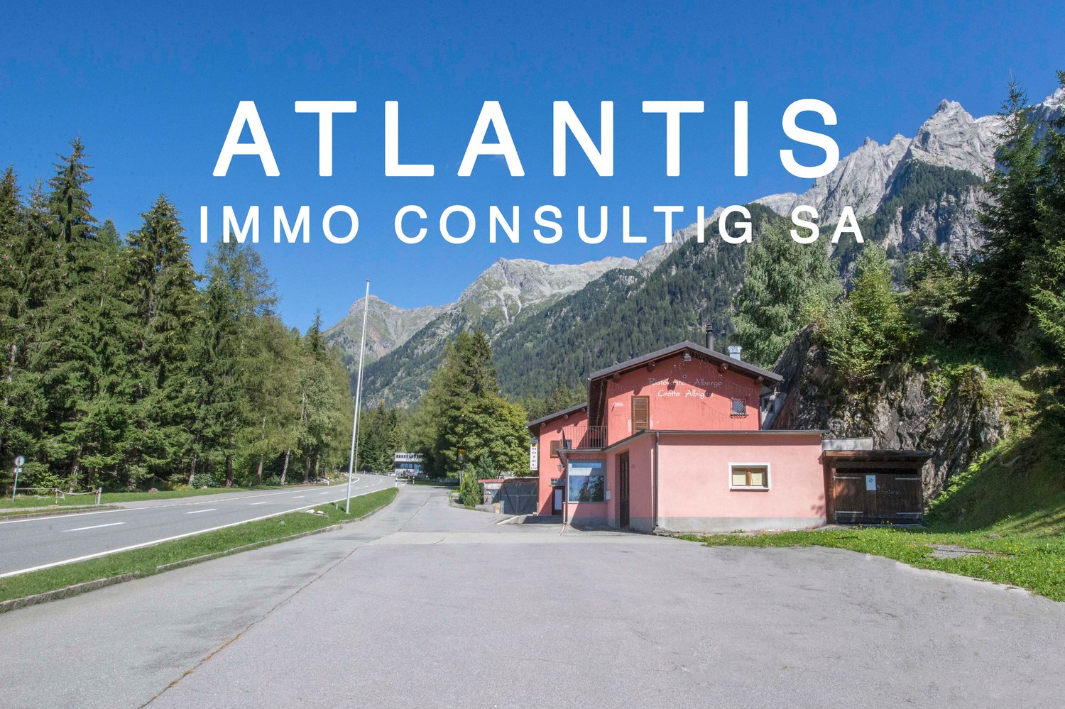 A red building with a small garden in front, beside a road with lush green trees on both sides and mountains in the background.