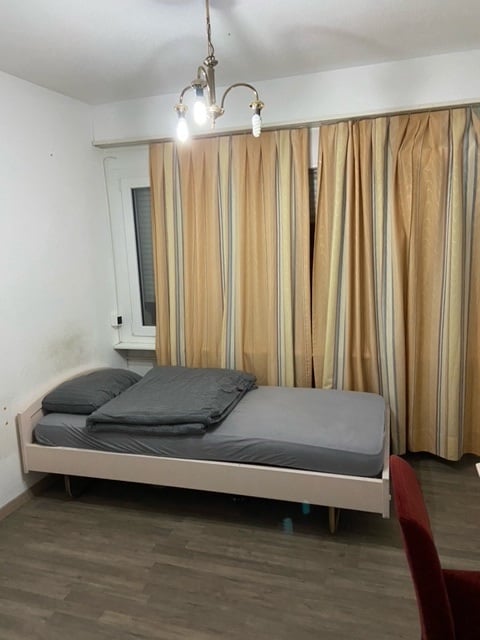 A bedroom with a single bed, wooden floor, beige walls, ceiling light, curtains, a chair, and a desk