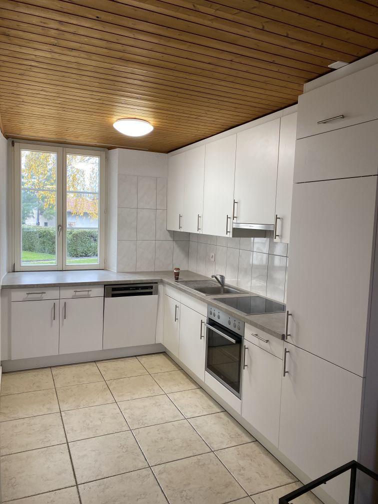 Modern kitchen with white cabinetry, stainless steel sink, tiled floor, dishwasher, washing machine, window with view to garden, built-in stove, oven, wooden ceiling