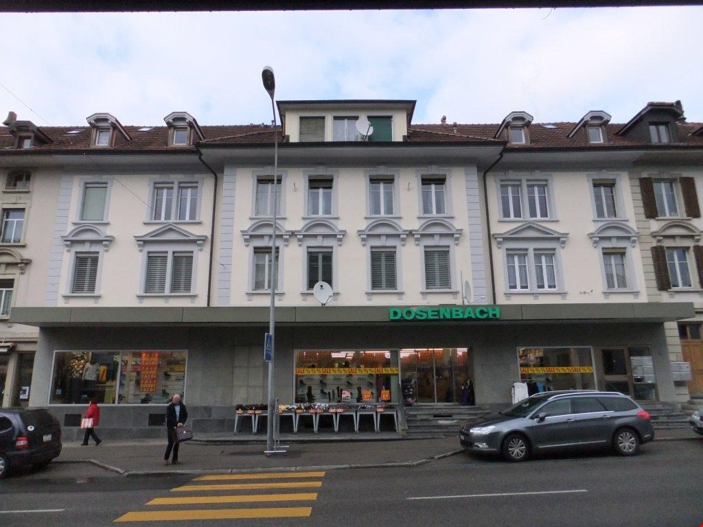 A multi-story building with a retail store called Dosenbach on the ground floor. The building has a traditional European architectural style with arched windows and decorative elements. There are people walking on the street in front of the building.