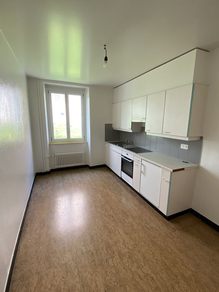 The image shows a modern kitchen with white cabinets, a tiled backsplash, and a window overlooking the exterior. The kitchen has a minimalist design with a clean and open layout. The floor appears to be made of a light-colored material, possibly linoleum