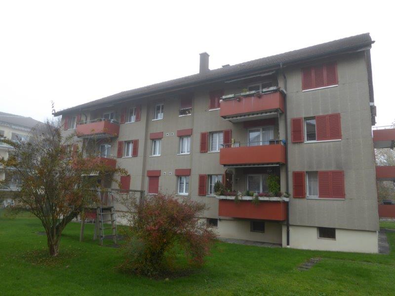 3-story apartment building with red and gray exterior, balconies on each unit, surrounded by grass and trees