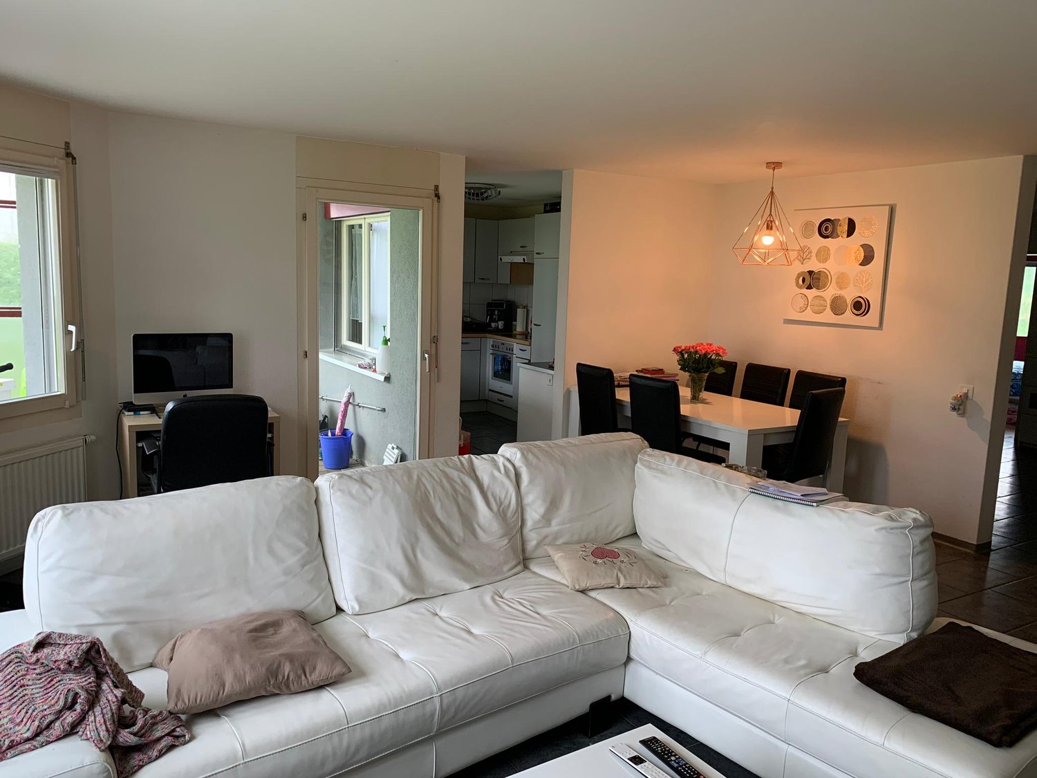 Spacious living room with white leather sectional sofa, dining table with black chairs, kitchen visible in the background, large window, decorative wall art, and pendant lighting fixture