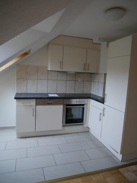 The image shows a kitchen area with white cabinets, a built-in oven, and tile flooring. The kitchen appears to be in a small apartment or studio space with slanted ceilings.