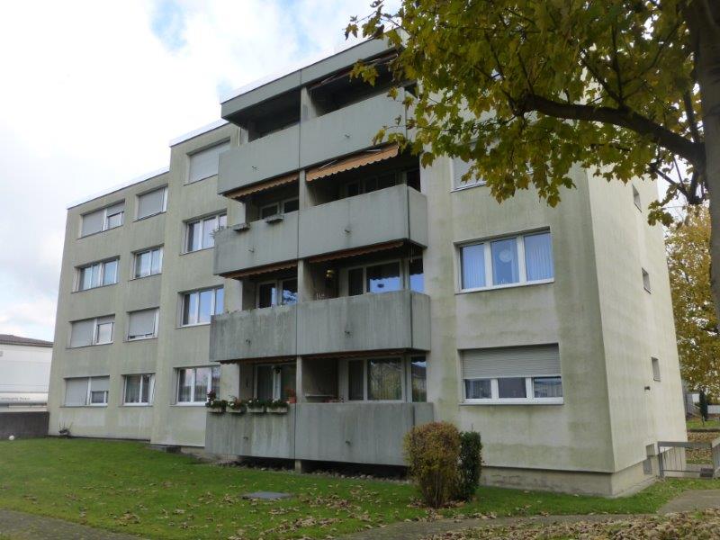 multi-story building, gray exterior, multiple windows, several balconies, fenced garden area, shrubs, trees