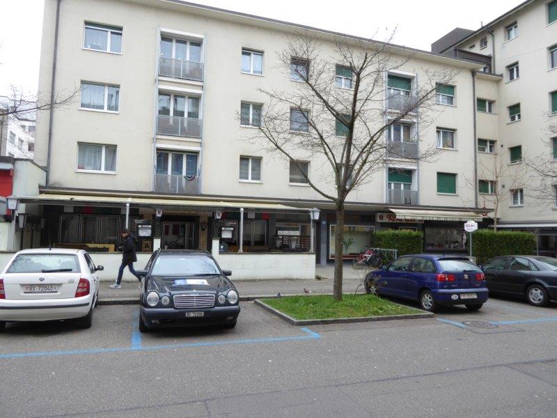 5-story white building with several windows, car parked in front of building, ground floor has windows with a bar