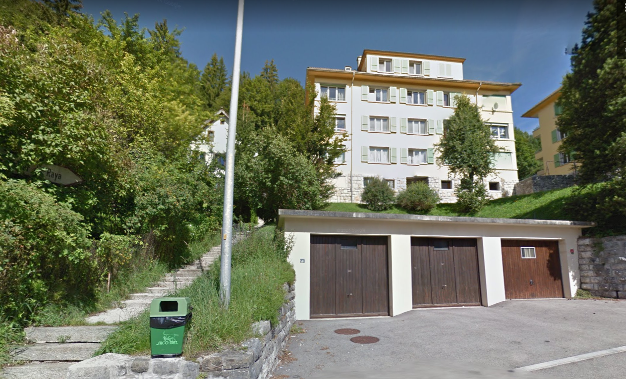 A multi-story apartment building with a garage on the ground level, surrounded by trees and greenery in a hilly, wooded area.