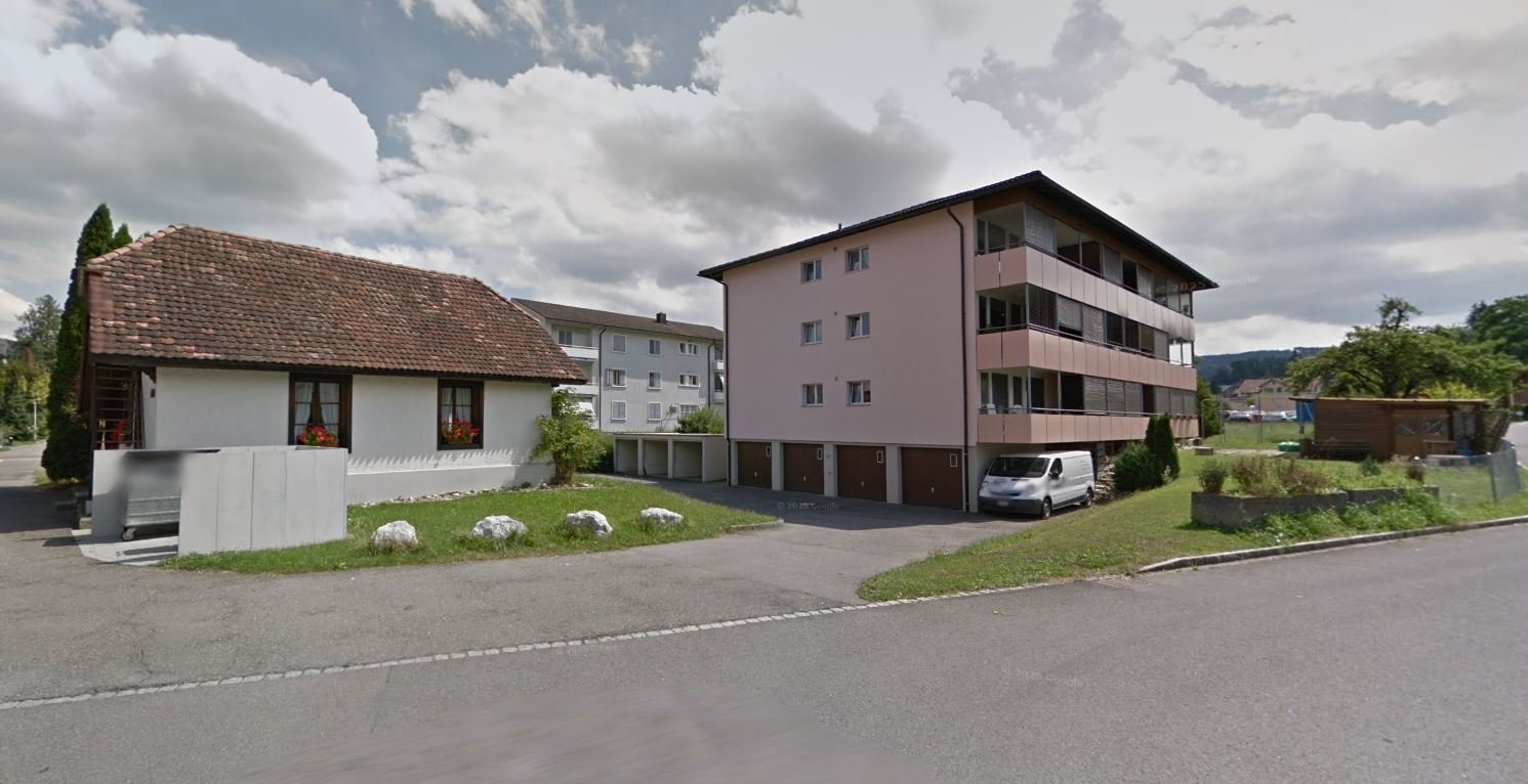 A raised ground floor white house, a garage, and a pink apartment building, all behind a road and grass.