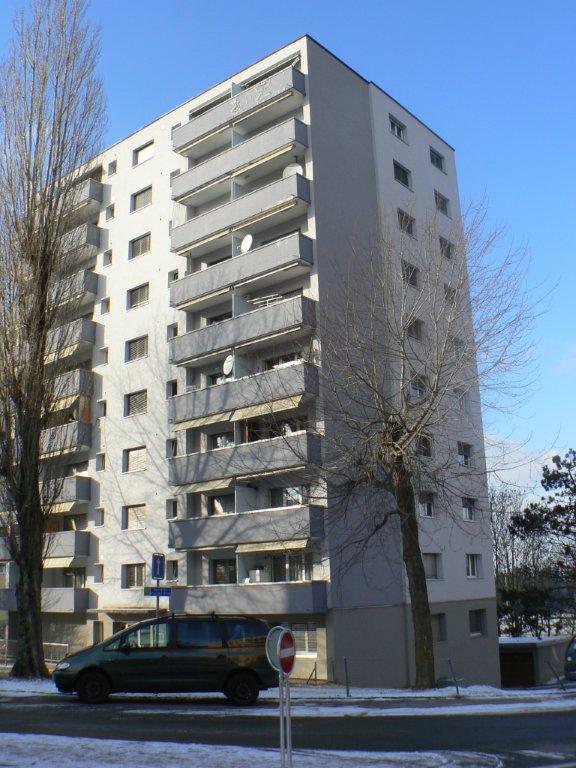 Multi-story apartment building with balconies, located in a snowy urban setting with a car parked in front.