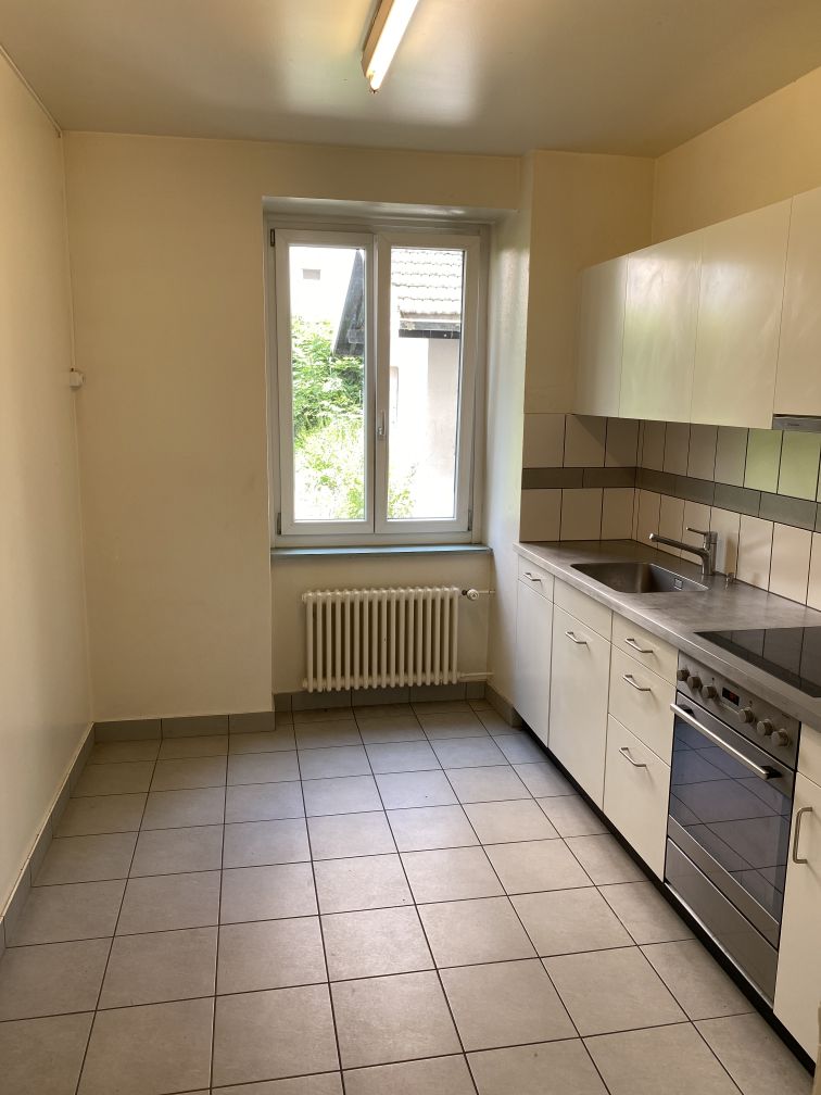 Empty kitchen with white tile flooring, white cabinets, sink, stove, window with a white radiator underneath