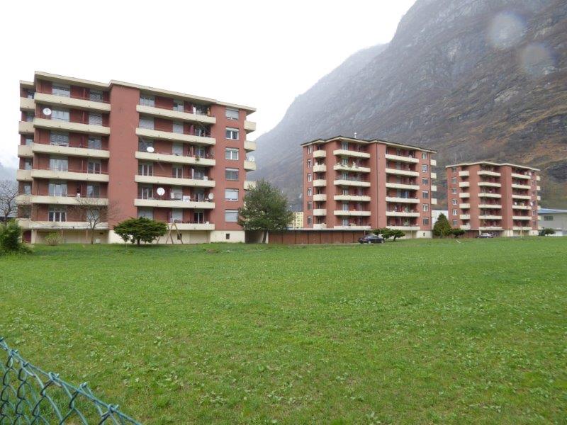 Multiple apartment buildings in the background, empty field, greenery, mountain, cars parked in front