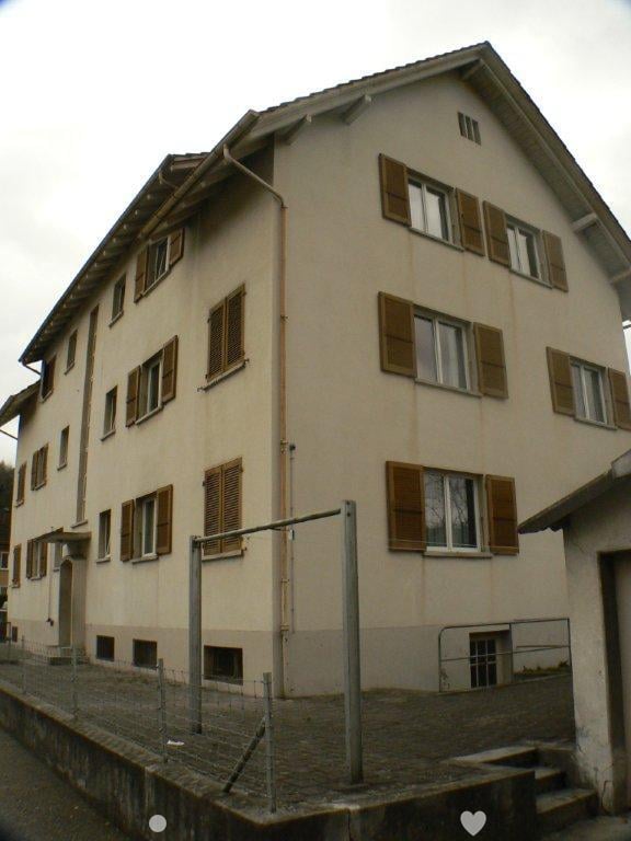 Multi-story residential building, light beige walls, wooden shutters, steep roof, fenced ground