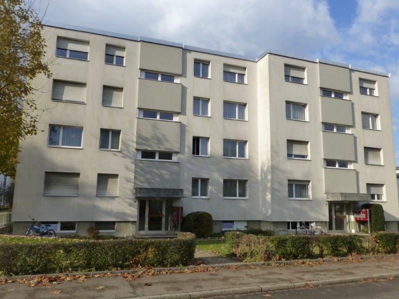 Modern residential building with concrete exterior, multiple windows, bicycles parked in front, bushes and trees in the foreground.