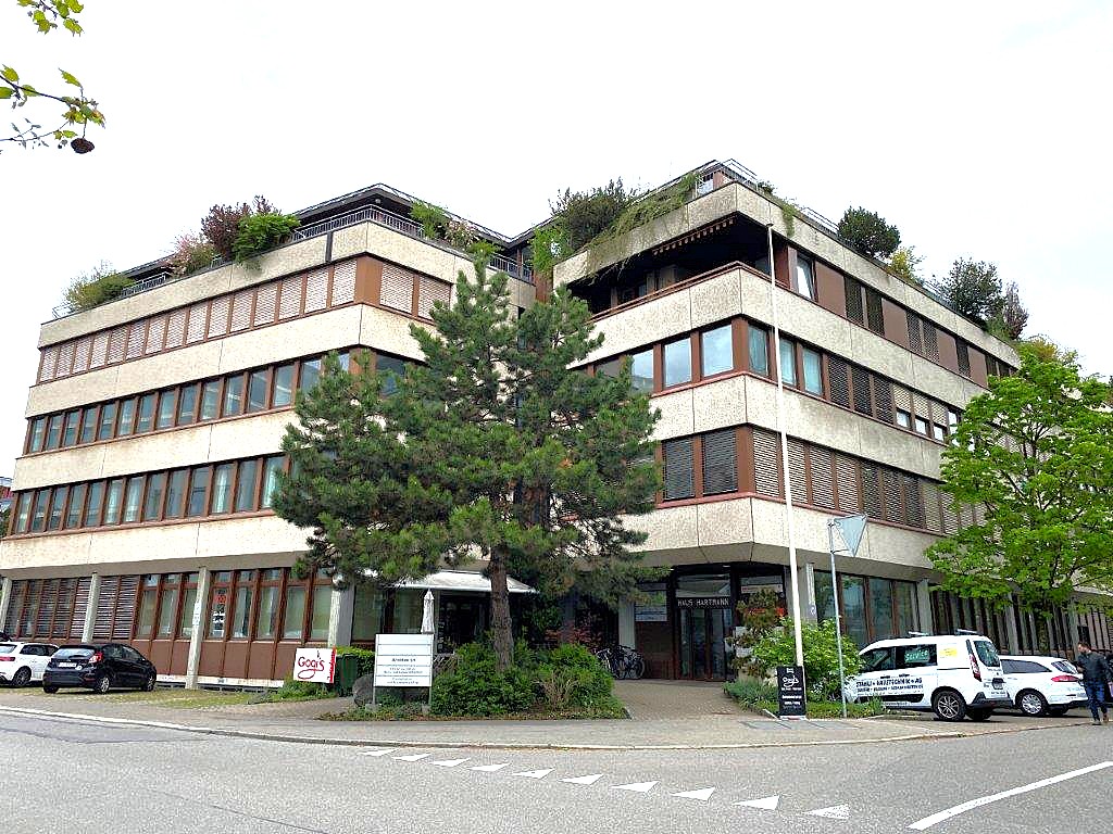 multi-story building, multiple windows, brown trim, parking area, green trees, and plants