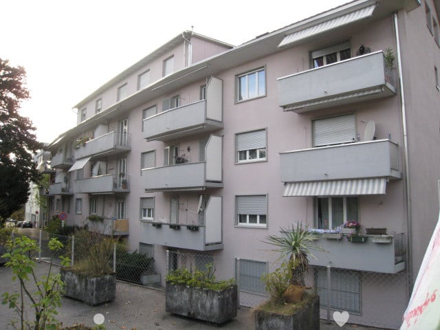 Multi-story apartment building with balconies, gray exterior, and landscaping around the building.