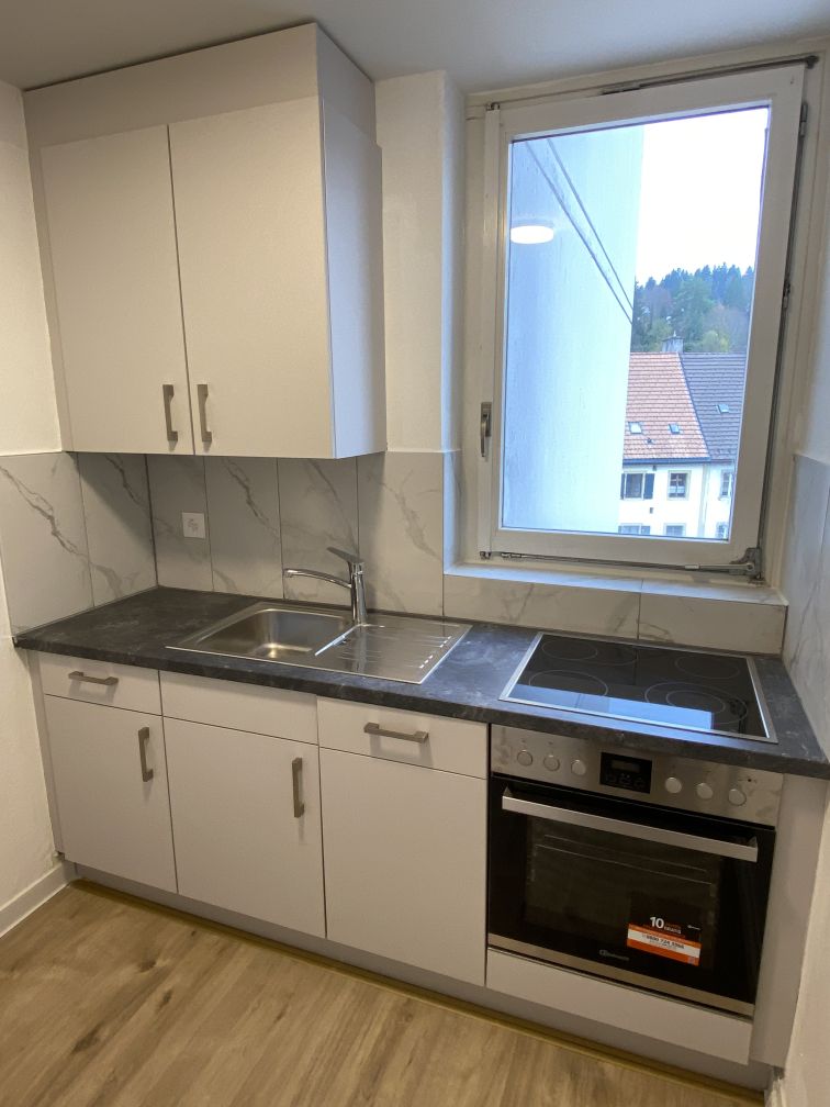 Modern kitchen with white cabinets, marble-like countertops, stainless steel sink, and built-in dishwasher. Large window overlooking the exterior of the building.