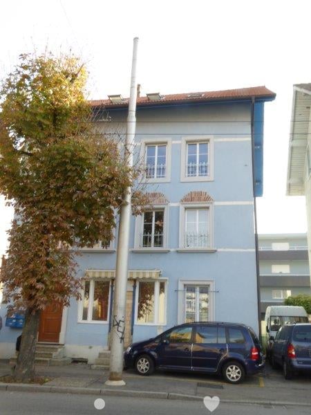 A three-story blue building with many windows and a brown door. Cars are parked on the street in front of the building.