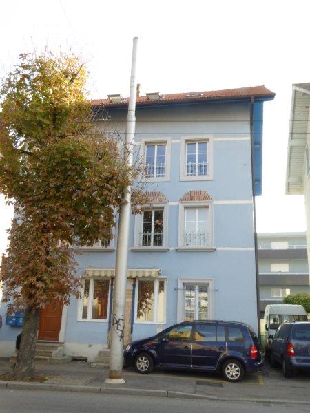 Blue three-story building, multiple windows, brown door, two cars parked, awnings, streetlight, tree