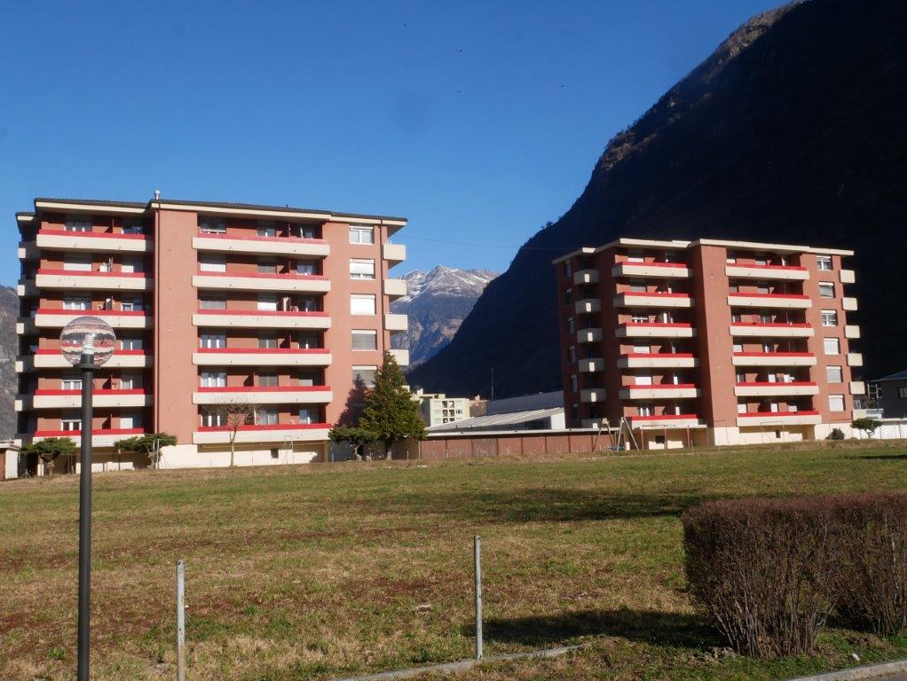 The image shows a multi-story apartment building with red and white exterior. The building has balconies on each floor and is situated in a mountainous area with a clear blue sky in the background.