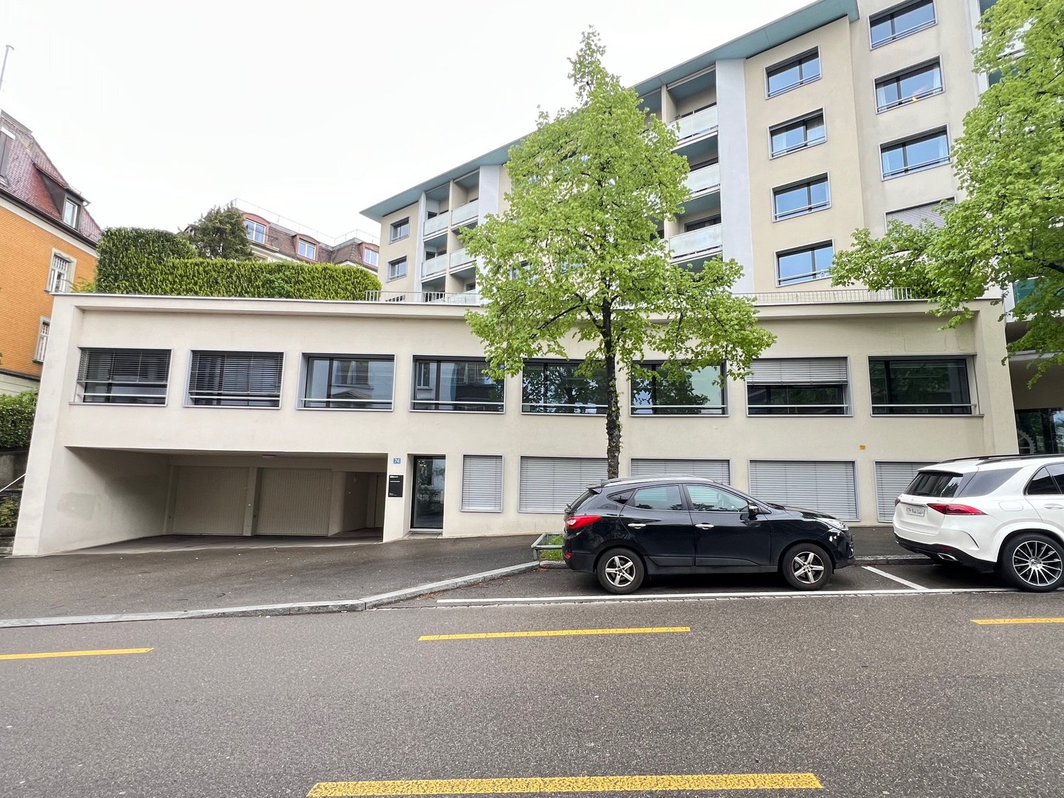 large apartment building, entrance garage with two cars parked, trees, multiple windows