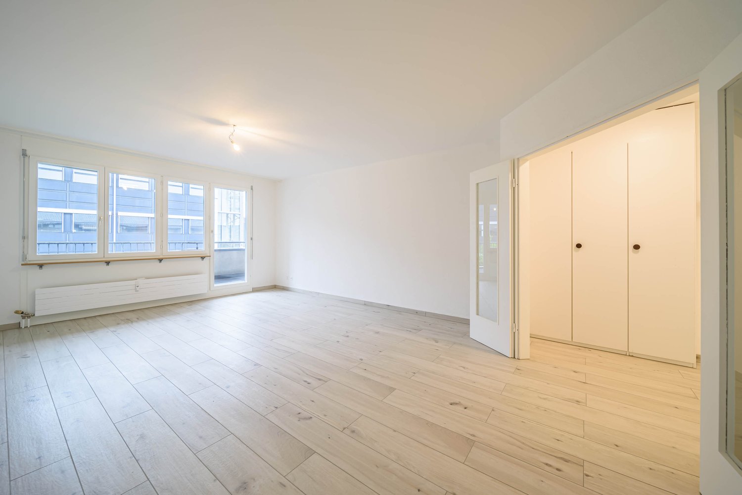 Empty room with wooden floor, white walls, three large windows, a radiator, a glass door and open closet