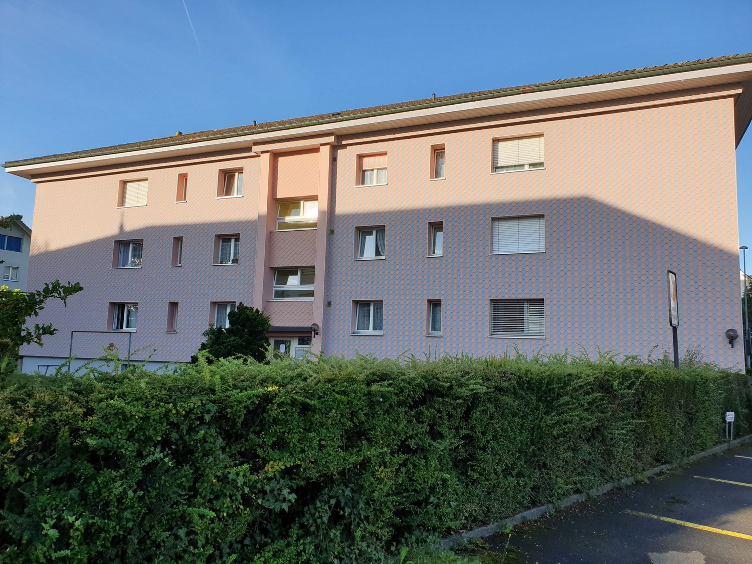 3-story apartment building, pink exterior with hexagonal tiles, windows with white shutters, front hedge, parking lot