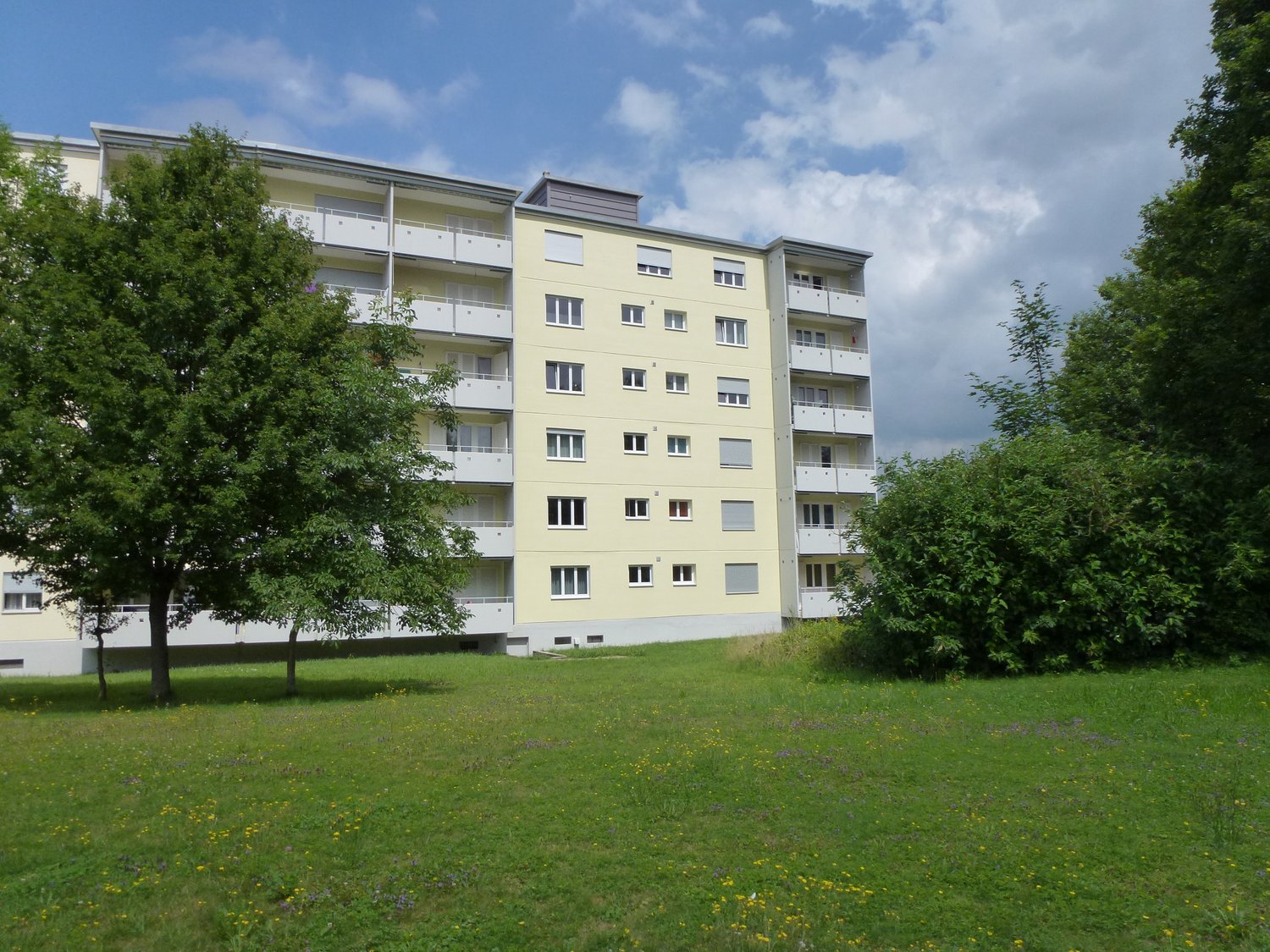 4 story building, yellow exterior, balcony on each floor, green lawn, trees