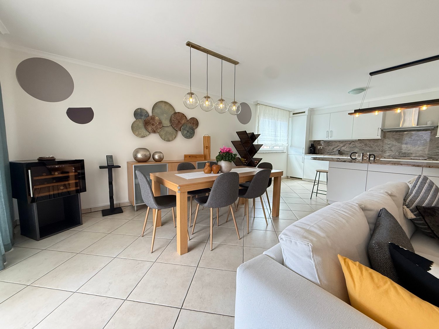 Dining room with a wooden table, gray chairs, a white couch with pillows, a black cabinet, a wine cooler, a kitchen with white cabinets, a countertop, and a hanging lamp.
