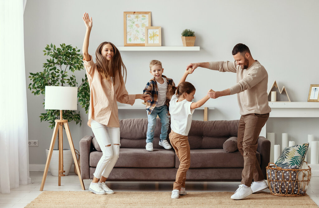 family dancing in living room, brown couch, wooden floor, shelf, basket, plant, lamp, candles