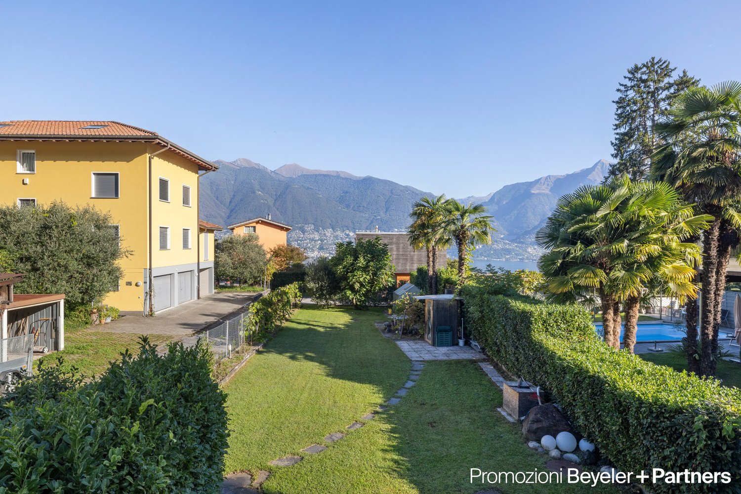 House with swimming pool and green gardens, yellow facade, mountains in the background.