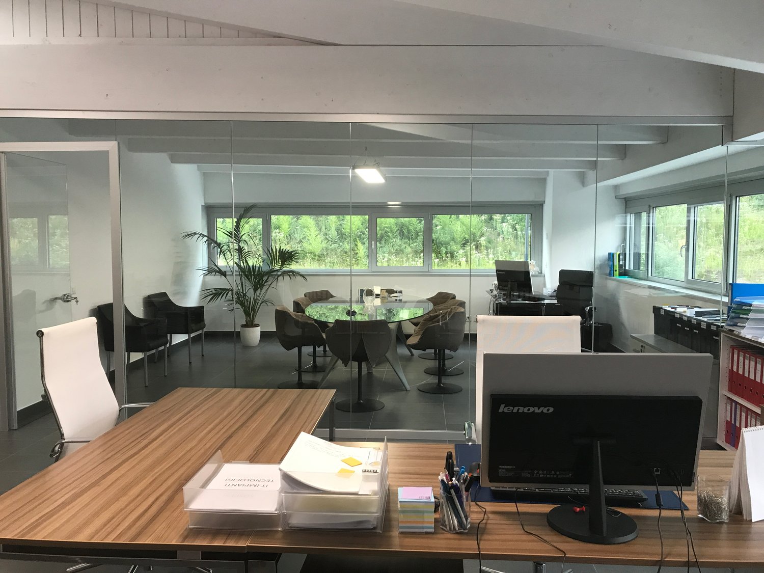 The image shows a modern and spacious office space with large windows providing a view of the surrounding greenery. The office features a glass-enclosed meeting area with a round table and comfortable chairs. There is also a desk with a Lenovo computer an