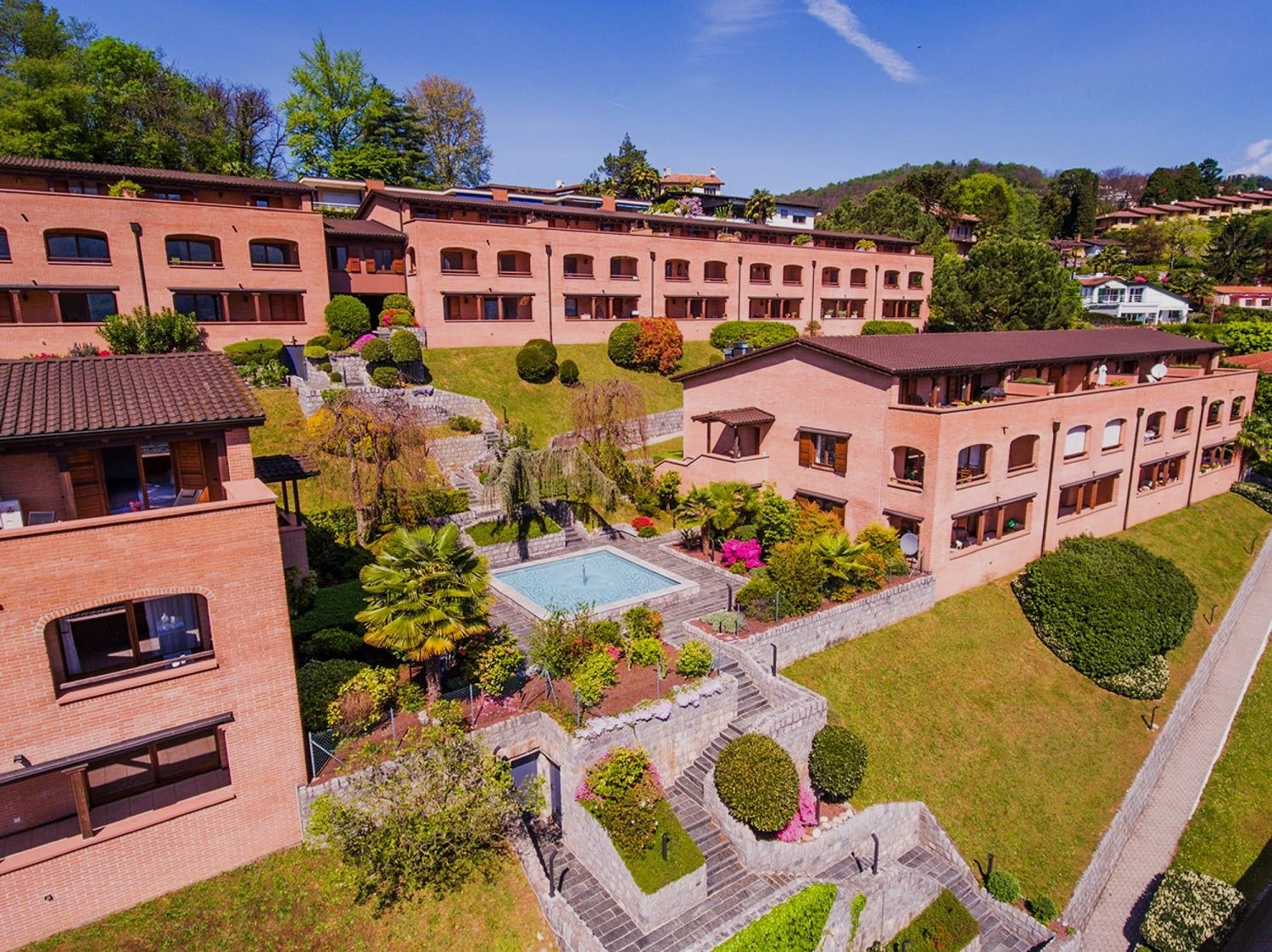 Multi-story brick building with red tile roofs, surrounded by lush gardens, landscaping, and a swimming pool. The building has balconies and terraces overlooking the grounds.