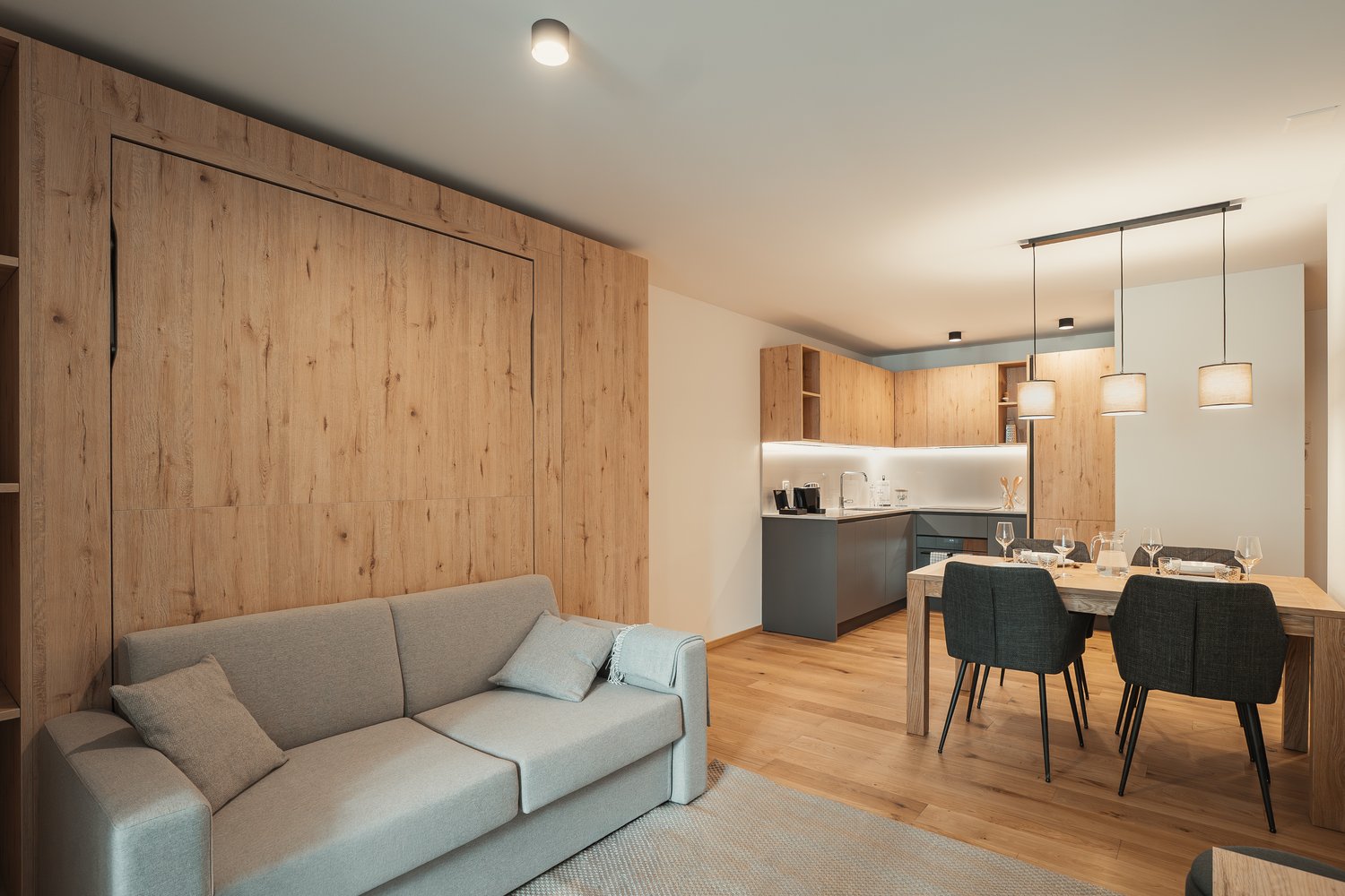 Open-plan living and dining area with a gray sofa, dining table, and wooden flooring. The kitchen is partially visible with a sink and cabinets.