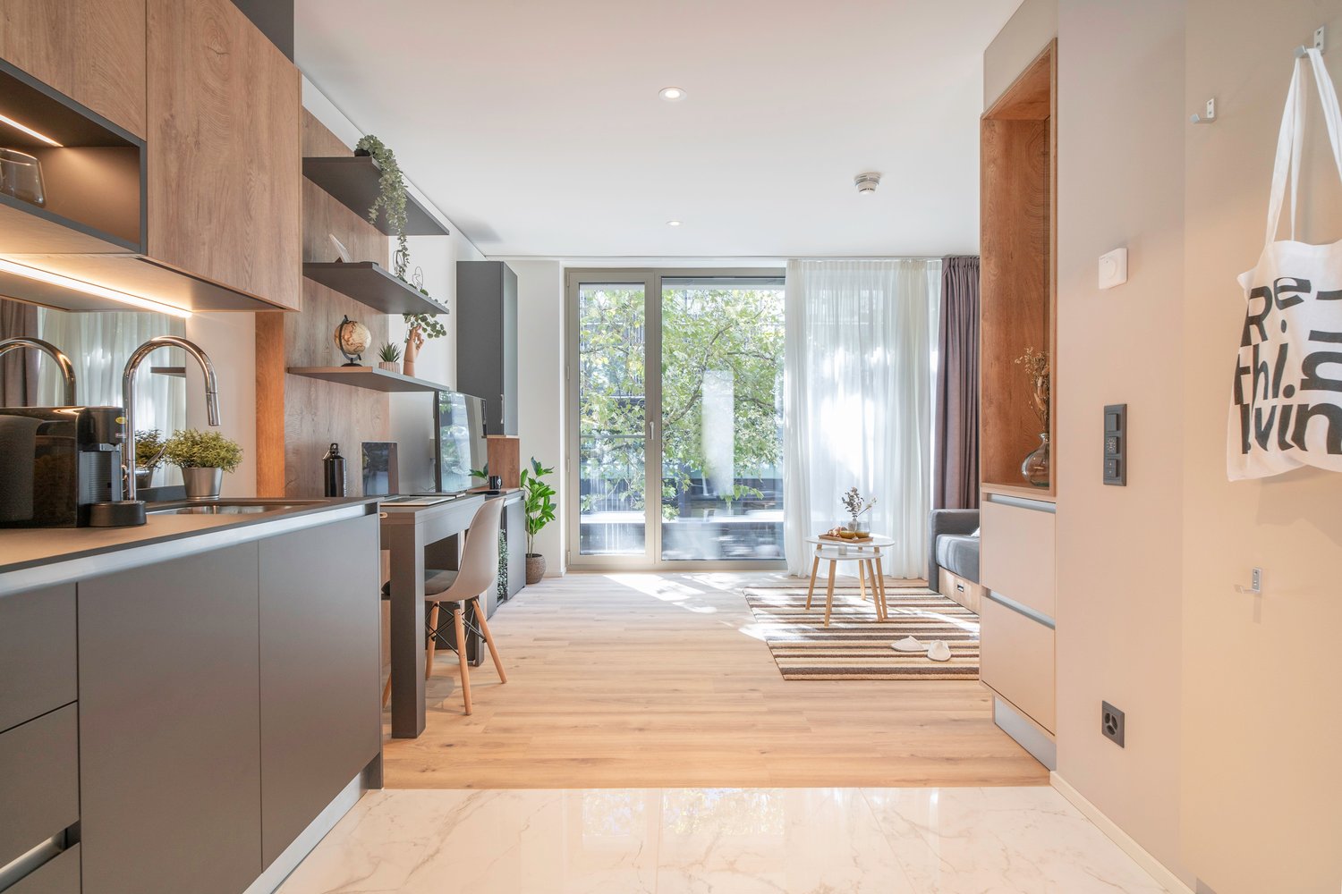 The image shows an open concept living space with a kitchen, dining area, and living room. The kitchen features modern gray cabinets, wooden shelves, and stainless steel appliances. The dining area has a small table and chairs, and the living room has a c