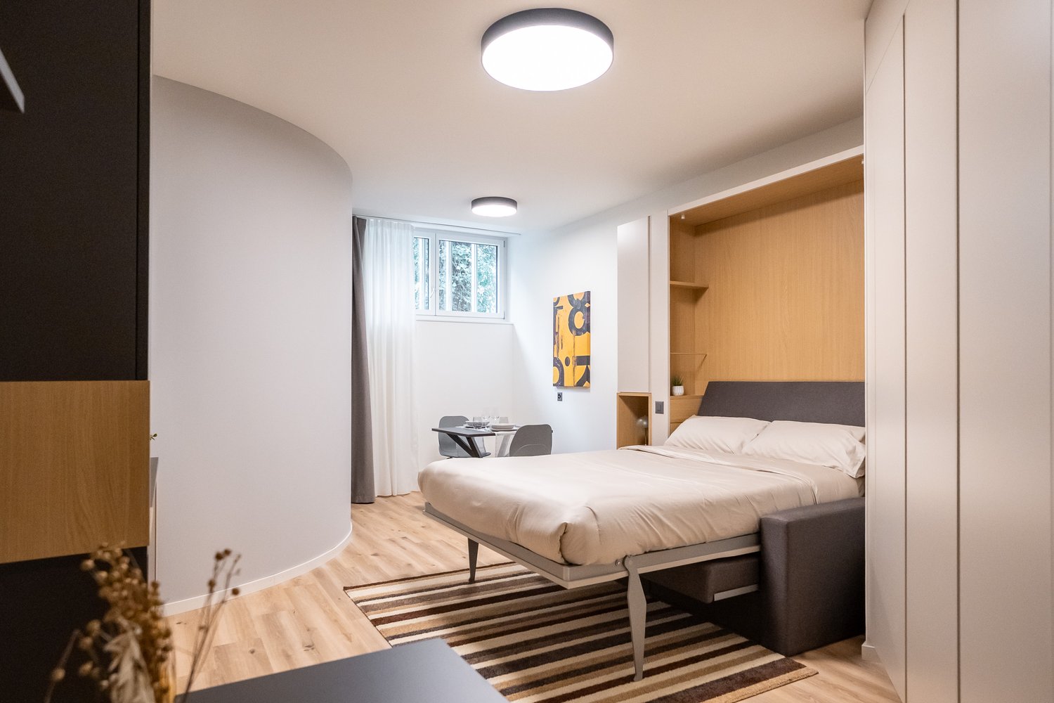 Spacious bedroom with hardwood flooring, white walls, large window, modern bed frame, built-in wardrobe, and a desk area