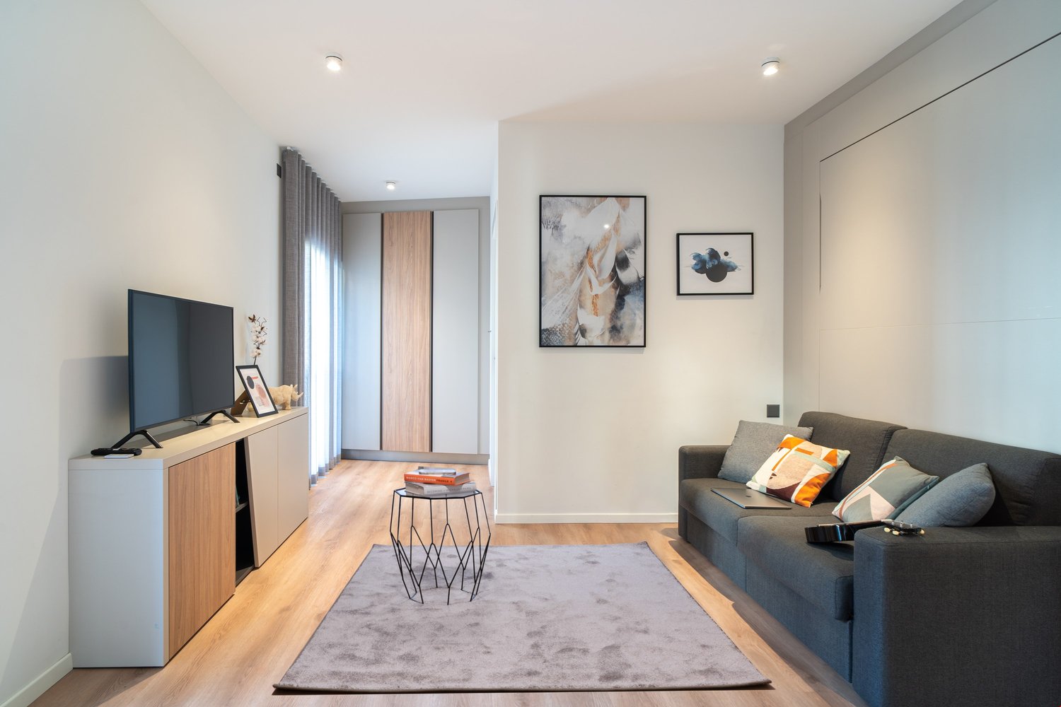 Spacious living room with hardwood floors, white walls, large windows, gray sofa, TV stand, and decorative artwork on the walls. The room also features a sliding door leading to a balcony or terrace.