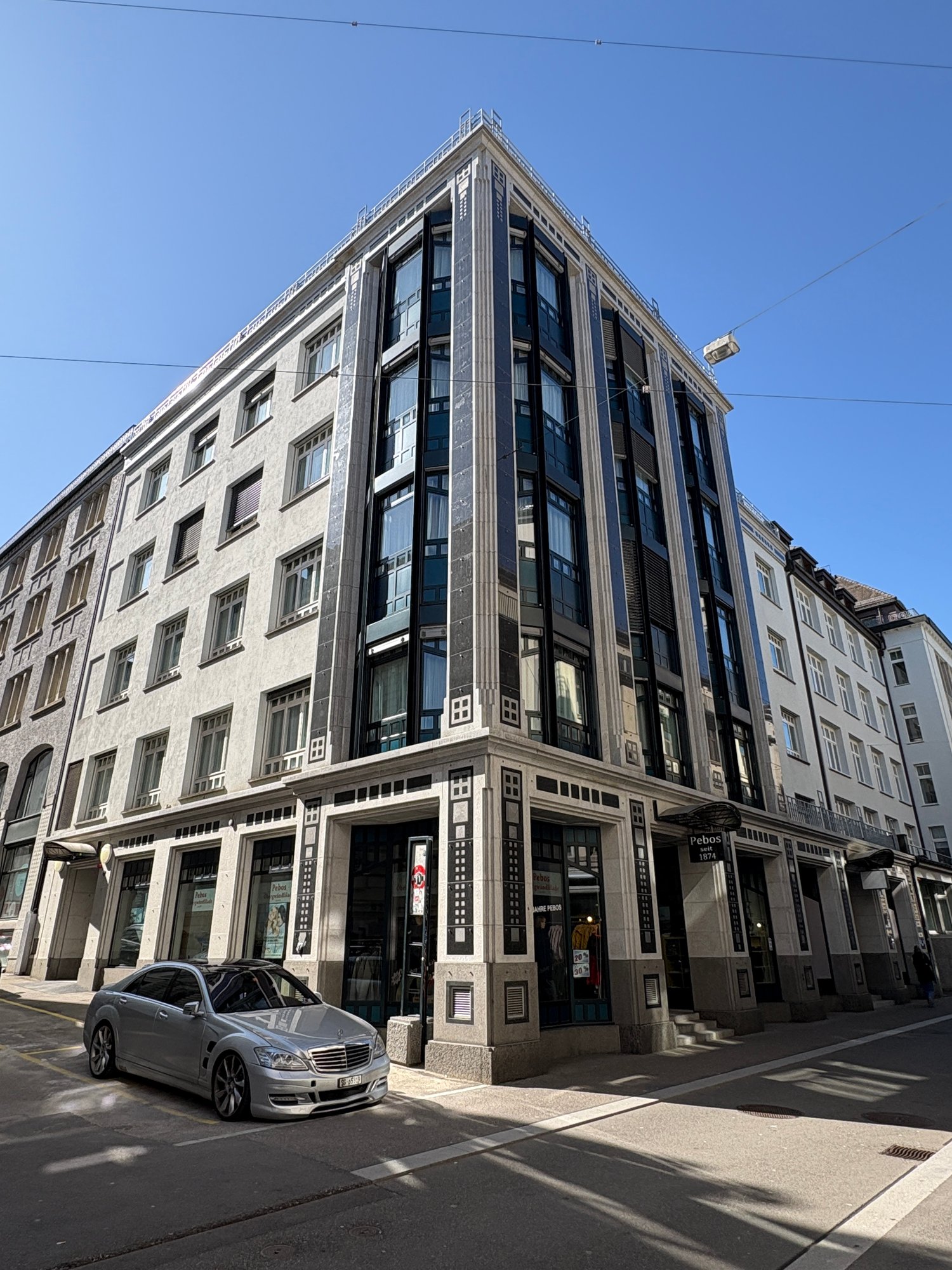 A multi-story commercial building with a distinctive architectural style, featuring a mix of glass and stone elements. The building has a prominent corner design with a recessed entrance and large windows. There is a parked car visible in the foreground.