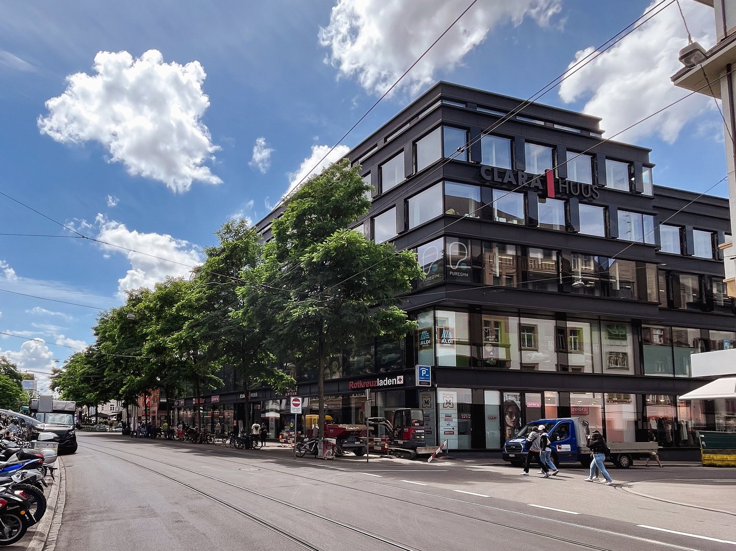 A multi-story commercial building with a modern, black and glass facade. The building has a sign that says 'Clara Haus' and there are various shops and businesses visible on the ground floor. There are people walking on the street in front of the building