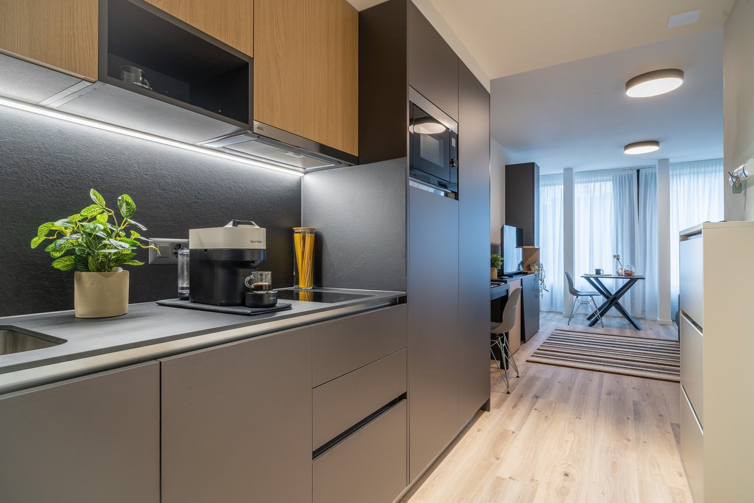 Modern kitchen with wood-tone cabinets, stainless steel appliances including a dishwasher, and a potted plant on the counter. The kitchen opens up to a dining area with a glass table and chairs.