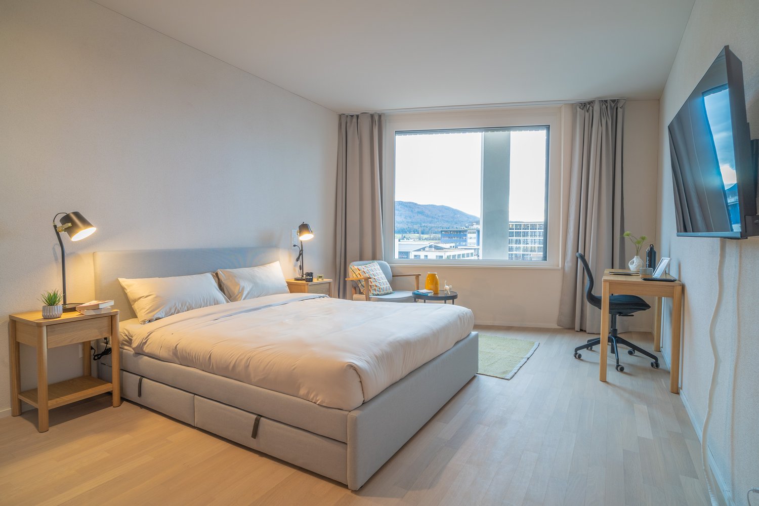 Spacious bedroom with large windows offering a scenic view, hardwood flooring, a king-size bed with storage drawers, two bedside tables with lamps, a desk with a chair, and a flat-screen TV mounted on the wall.