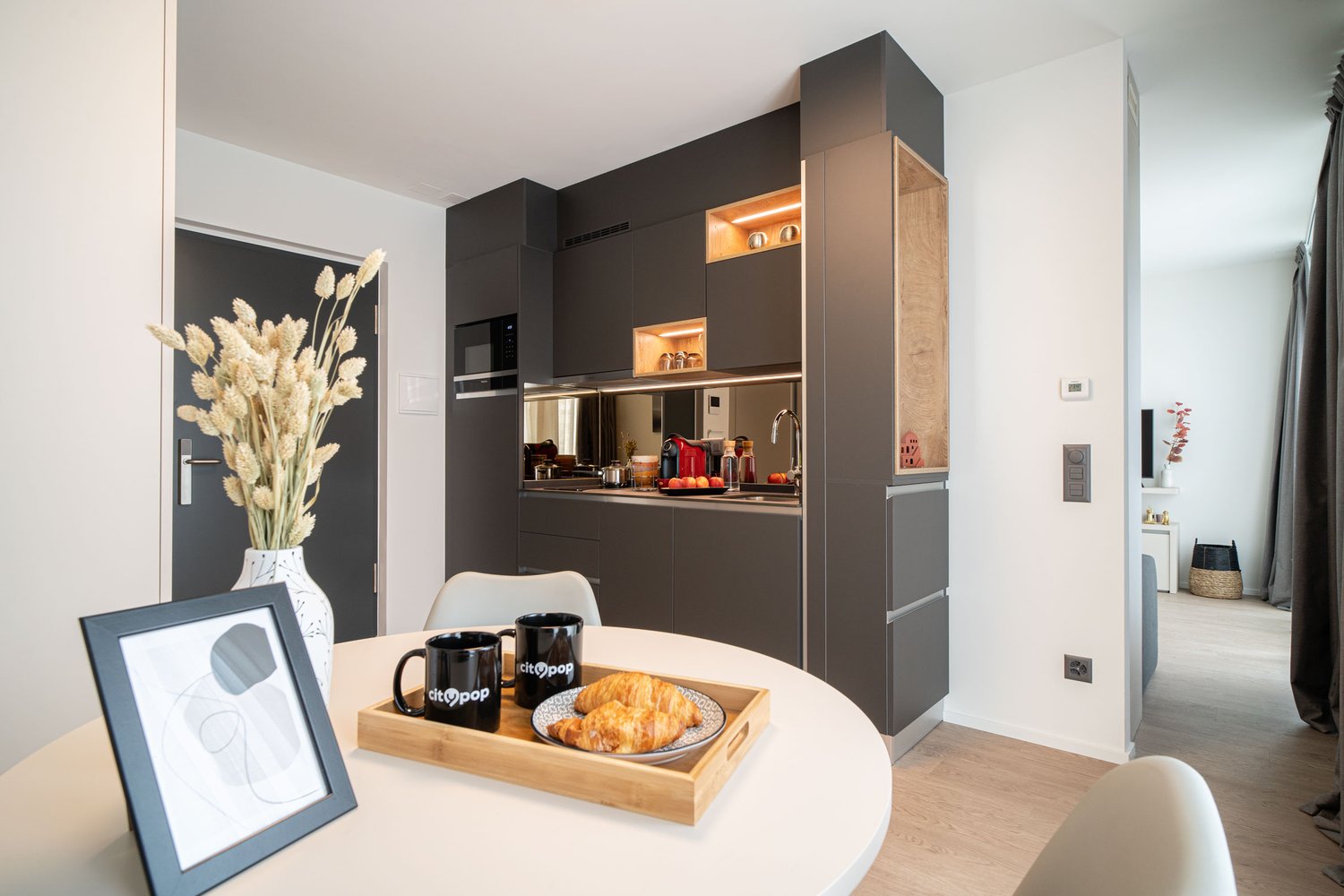 Modern kitchen with dark cabinets, built-in appliances, and a dining area with a round table and chairs. The kitchen features a dishwasher and is decorated with a vase of dried flowers and framed artwork on the wall.