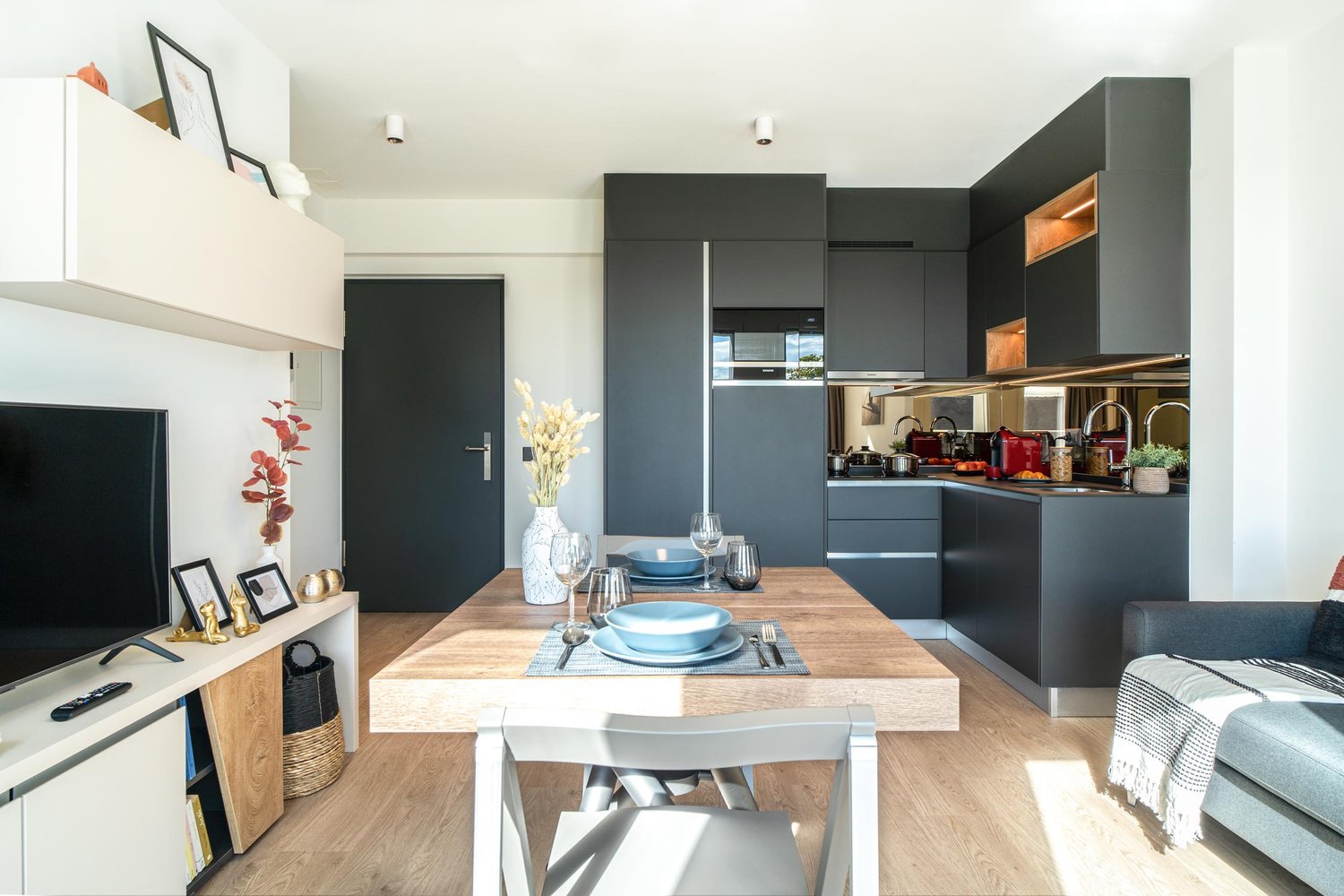 The image shows an open-plan living room and kitchen area with a large wooden dining table, black kitchen cabinets and appliances, and a cozy living space with shelving and decor. The kitchen appears to have a dishwasher visible as one of the appliances.