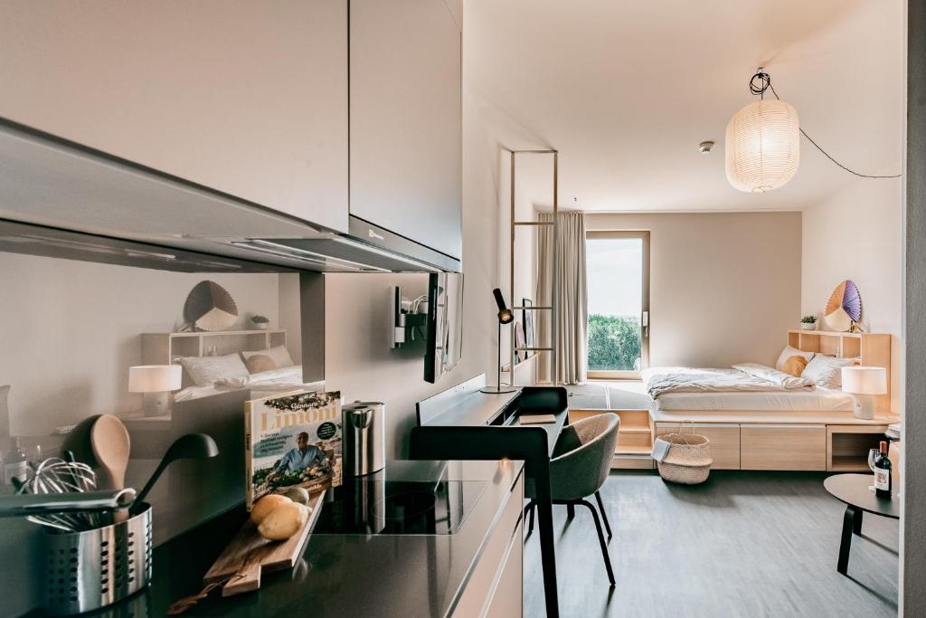 Open-plan studio apartment with kitchen, dining area, and bedroom. The kitchen features modern appliances including a dishwasher. The living space has a bed, side tables, and a pendant light fixture. The overall decor is minimalist and neutral-toned.
