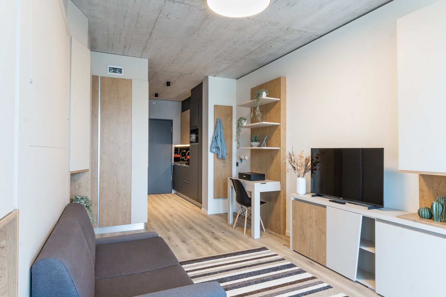 Open-concept studio apartment with concrete ceiling, wood-paneled walls, hardwood flooring, sofa, TV stand, desk, shelving unit, and a refrigerator visible in the kitchenette area.