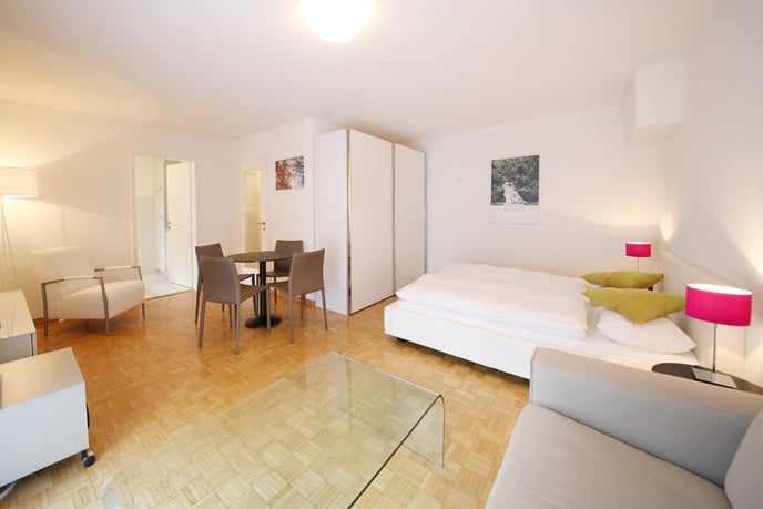 Open-plan studio apartment with hardwood floors, white walls, large windows, dining table, sofa, and a bed area with a wardrobe.