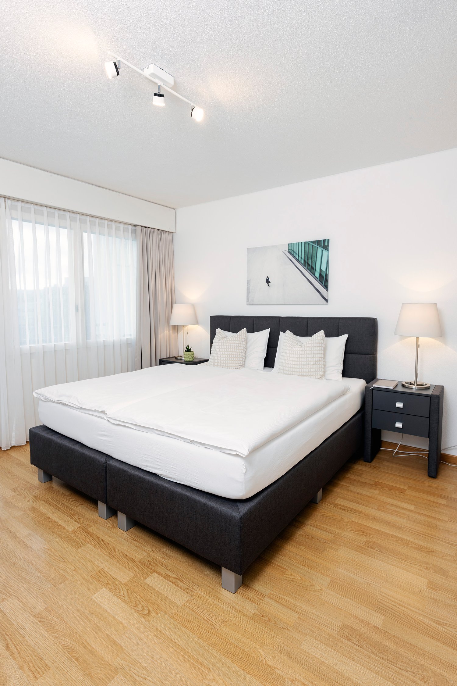 spacious bedroom with large bed, lamps, side tables, nightstands, white linens, framed photo, wooden floor, large window with white curtains