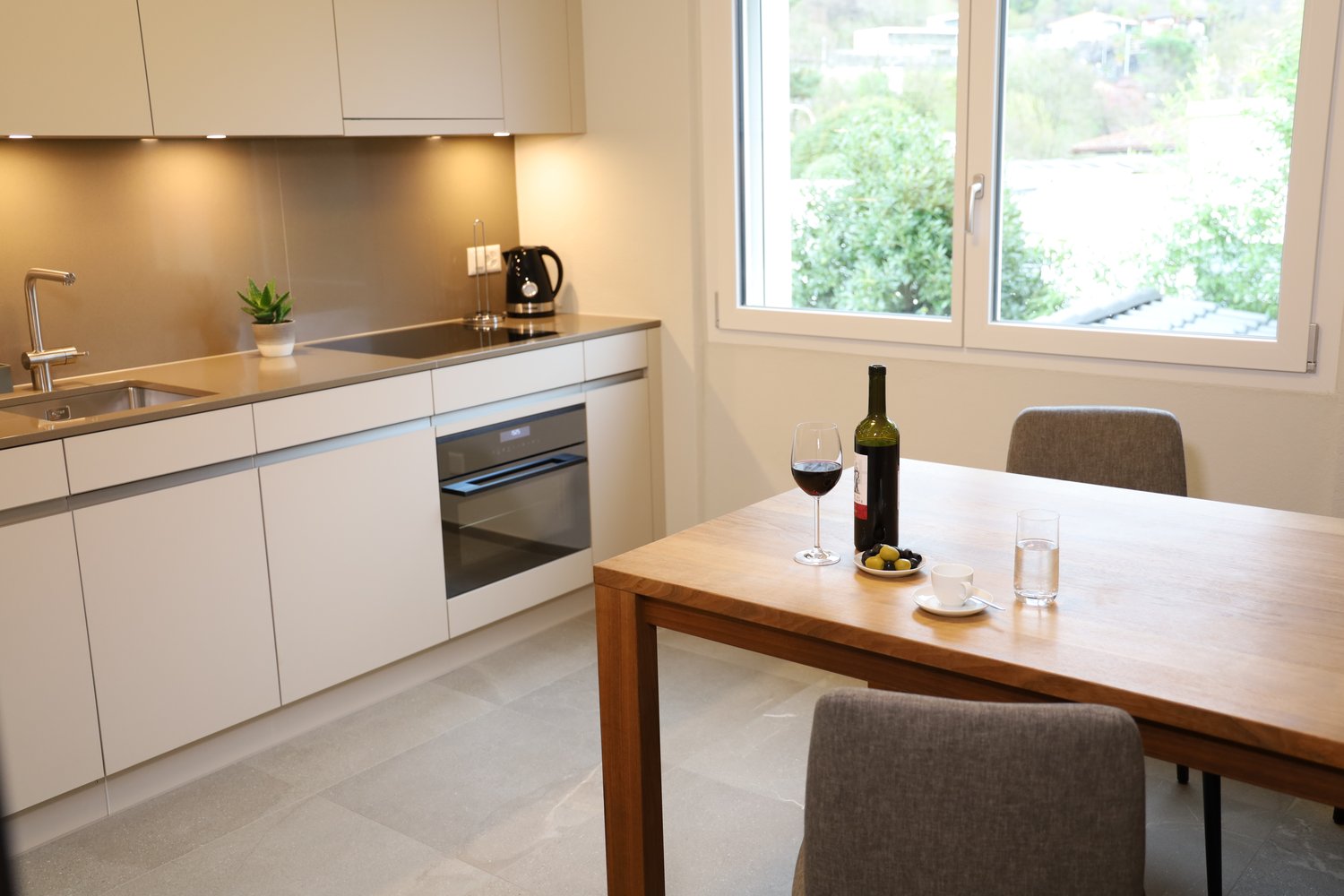 Modern kitchen with white cabinets, granite countertop, sink with faucet, electric stove, oven, wine glasses, wine bottle, plate, glass, coffee cup, grey chairs, large window
